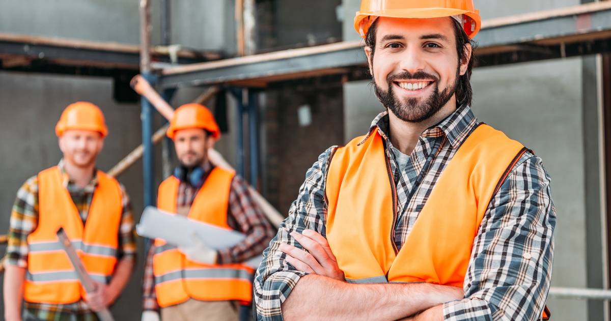 knappe bouwondernemer staat met zijn armen over elkaar op een bouwplaats met zijn collega's op de achtergrond